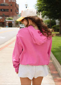 A woman seen from behind wearing a bright pink pullover hoodie, light gray shorts, a white baseball cap with a yellow brim, standing on a city sidewalk.