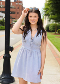 Woman in a light blue dress standing on a sidewalk with a building and greenery in the background