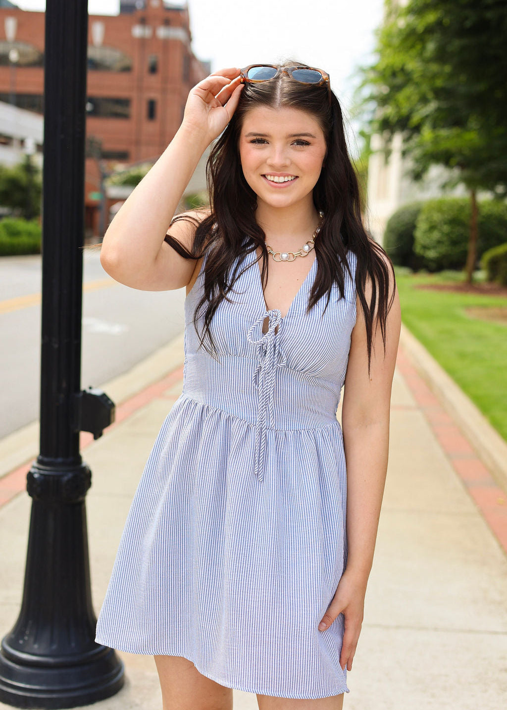 Woman in a light blue dress standing on a sidewalk with a building and greenery in the background