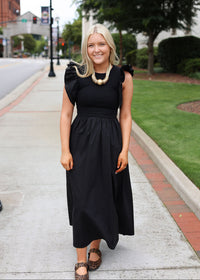 A woman stands on a city sidewalk wearing a black midi dress with puffed cap sleeves, a gathered waist, and a flared skirt. She is accessorized with a chunky gold statement necklace and leopard-print flats.