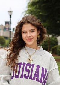 A woman wearing an oversized light gray sweatshirt with 'LOUISIANA' printed in purple across the chest, standing on a sidewalk outdoors.