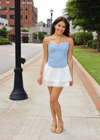 Woman in a light blue top and white skirt standing on a sidewalk with buildings and trees in the background.