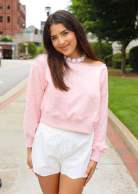 A woman standing outdoors on a sidewalk wearing a light pink off-the-shoulder long-sleeve top with white polka dots and white shorts.