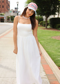 Woman in a white dress and striped cap standing on a sidewalk with greenery in the background
