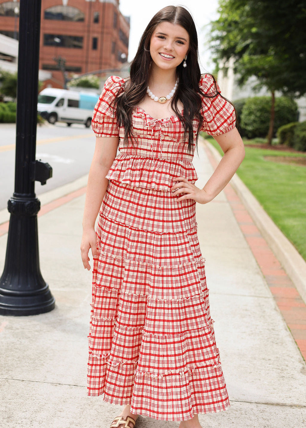 Woman in a red and white checkered dress standing on a sidewalk with buildings and trees in the background.