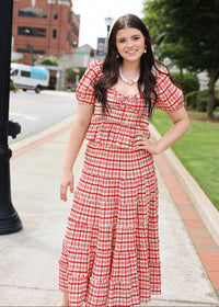 Woman in a red and white checkered dress standing on a sidewalk with buildings and trees in the background.