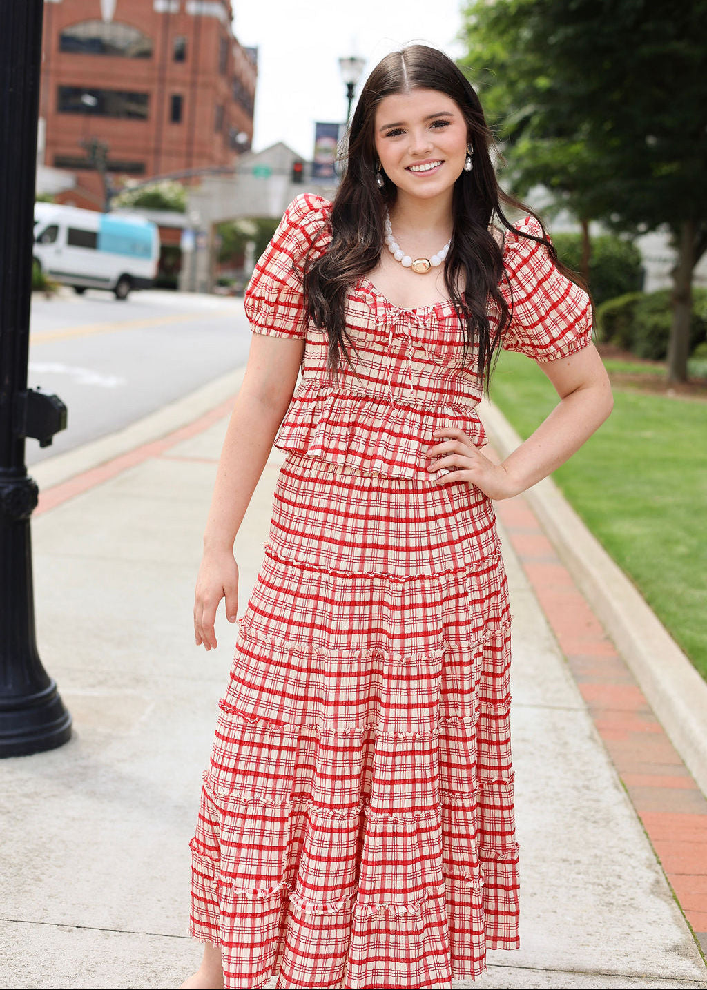 Woman in a red and white checkered dress standing on a sidewalk with buildings and trees in the background.