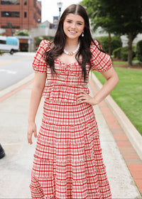 Woman in a red checkered dress standing on a sidewalk with buildings and trees in the background