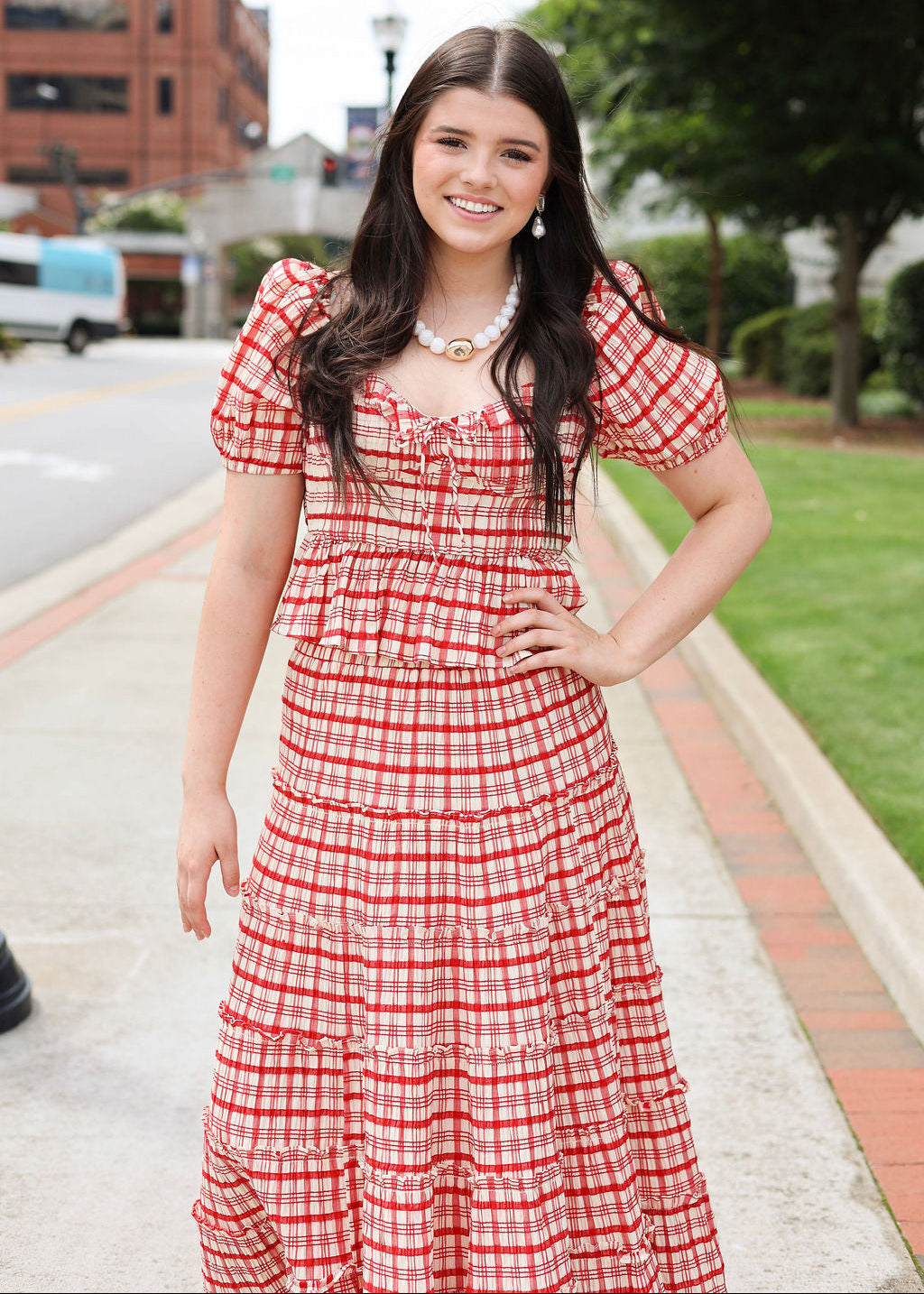 Woman in a red checkered dress standing on a sidewalk with buildings and trees in the background