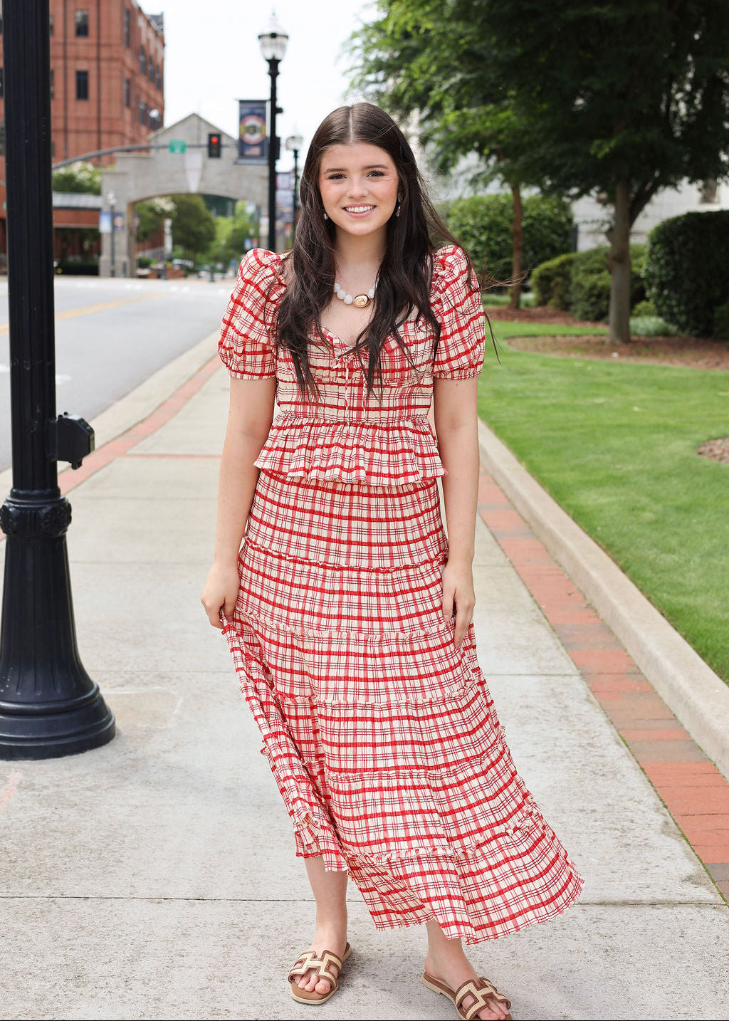Woman in a red and white checkered dress standing on a sidewalk with greenery and buildings in the background.