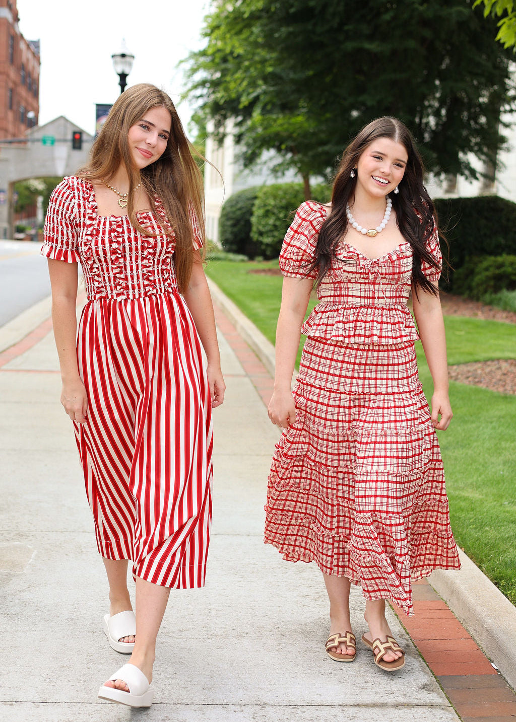 Two women wearing red and white dresses standing on a sidewalk with greenery in the background.